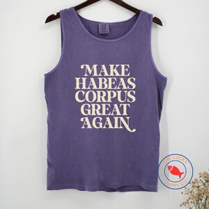 May include: A purple sleeveless tank top featuring the cream-colored text "MAKE HABEAS CORPUS GREAT AGAIN" printed on the front. The garment has a crew neckline and is displayed on a hanger. A circular logo with a red fish is visible in the corner.