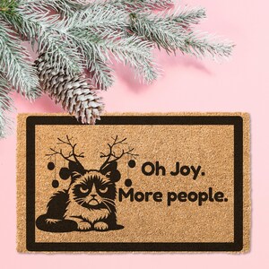 May include: A brown coir doormat with a black border and a black silhouette of a cat wearing reindeer antlers. The text "Oh Joy. More people." is printed on the doormat.