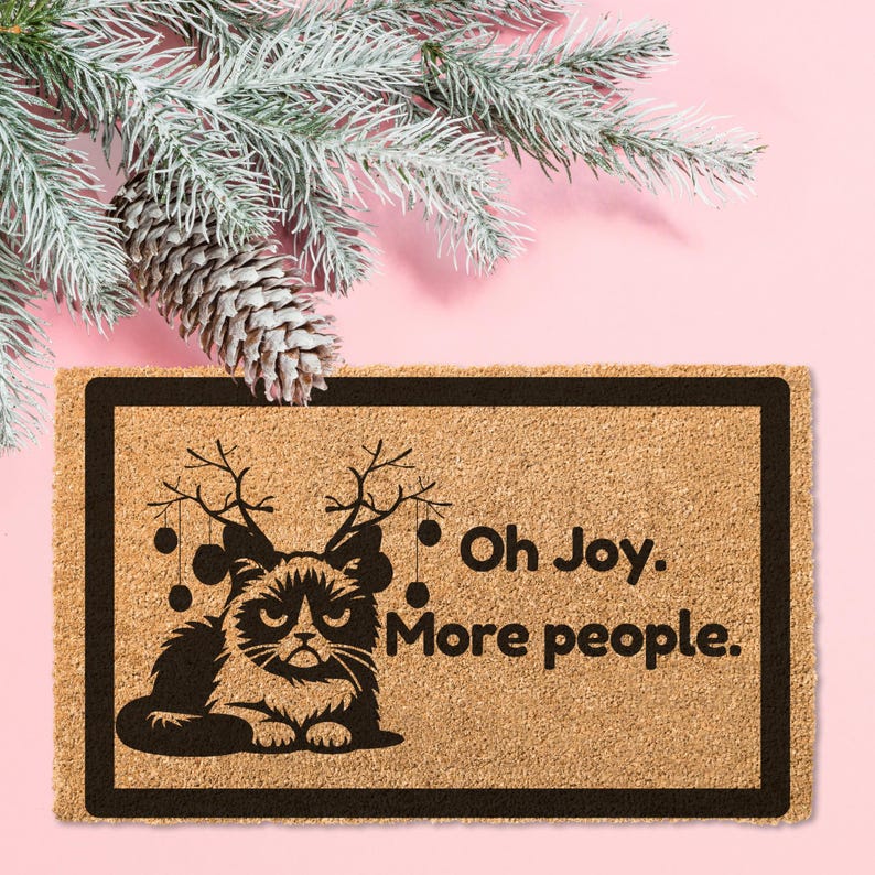 May include: A brown coir doormat with a black border and a black silhouette of a cat wearing reindeer antlers. The text "Oh Joy. More people." is printed on the doormat.