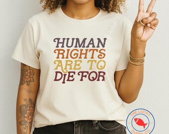 May include: A cream-colored short-sleeve t-shirt featuring the text "HUMAN RIGHTS ARE TO DIE FOR" printed in a stacked, multi-colored retro font. The letters transition from dark purple to orange and yellow across the four lines of text.