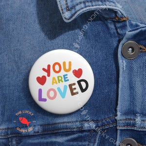 May include: A white button pin with the words "YOU ARE LOVED" in colorful letters and two red hearts. The pin is set against a blue denim jacket. The Lucky Guppy Co. logo is visible at the bottom.