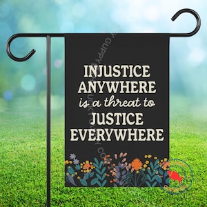 May include: A black garden flag with the quote "INJUSTICE ANYWHERE is a threat to JUSTICE EVERYWHERE" in white text. The bottom of the flag features a floral design with orange, yellow, and blue flowers. The flag is displayed on a black metal stand.