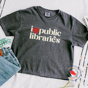 May include: Dark gray t-shirt with the text "i ❤️ public libraries" in a white, vintage-style font. The heart symbol is red. The shirt is short-sleeved and appears to be made of a soft, comfortable material. A pair of blue jeans is folded next to the shirt.