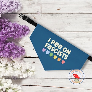 May include: A blue dog bandana with the text "i Pee ON Fascists" in white, with colorful heart graphics. The bandana has a black adjustable strap and buckle. The Lucky Guppy Co. logo is in the bottom right corner.