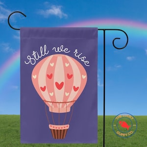 May include: A garden flag with a purple background features a hot air balloon design with pink and white stripes and red heart accents. The words "still we rise" are written in white script above the balloon. A rainbow is visible in the background.