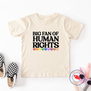 May include: Cream-colored t-shirt with the text "BIG FAN OF HUMAN RIGHTS" in black, arched lettering. Below the text are colorful heart graphics. The Lucky Guppy Co. logo is in the bottom right corner.