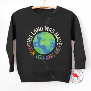 K&ouml;nnte beinhalten: Schwarzes Sweatshirt mit einer bunten Erdkugel-Grafik und dem Text "THIS LAND WAS MADE FOR YOU AND ME". Das Sweatshirt h&auml;ngt an einem Holzkleiderb&uuml;gel.