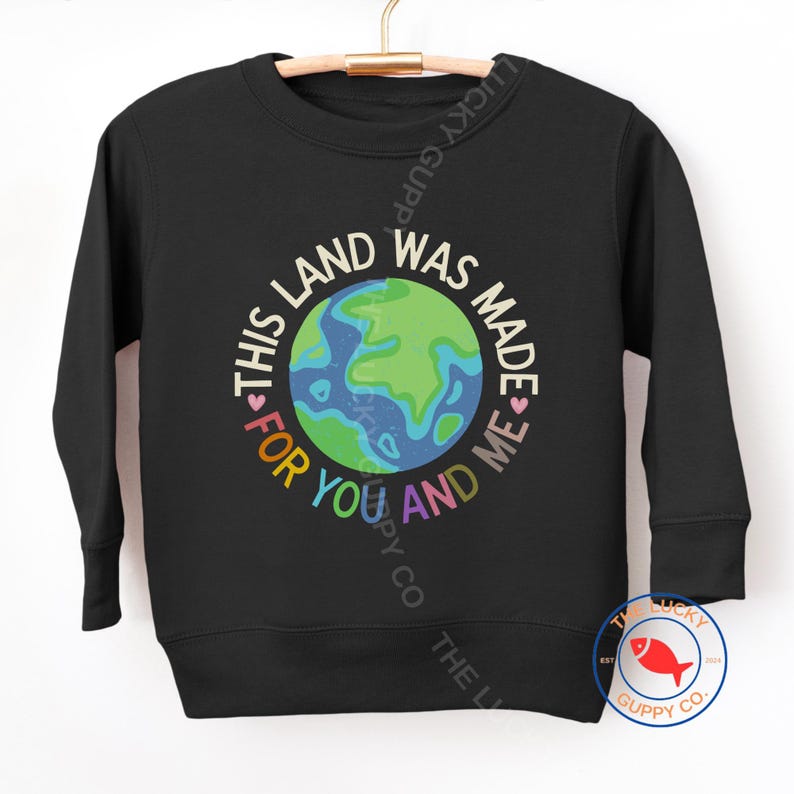 K&ouml;nnte beinhalten: Schwarzes Sweatshirt mit einer bunten Erdkugel-Grafik und dem Text "THIS LAND WAS MADE FOR YOU AND ME". Das Sweatshirt h&auml;ngt an einem Holzkleiderb&uuml;gel.