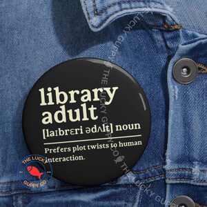 May include: A black button with the words "library adult" in white text, along with a definition. The button also features a red fish logo and the text "The Lucky Guppy Co."