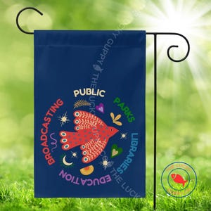 May include: A navy blue garden flag featuring a red bird design with the words "Public, Parks, Libraries, Education, Broadcasting" in a circular pattern. The flag is hanging on a black metal stand in a grassy outdoor setting.