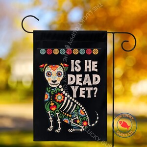 May include: A black garden flag featuring a colorful sugar skull-style dog illustration with floral patterns and skeletal details. The text "IS HE DEAD YET?" is printed in white capital letters next to the dog, with a row of small circular icons above.