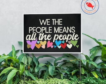 May include: A black sign with the text "WE THE PEOPLE MEANS all the people" in white and colorful hearts. The sign is displayed outdoors, surrounded by green plants and a white border. The Lucky Guppy Co. logo is in the top right corner.