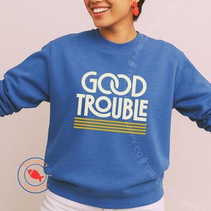 May include: A blue sweatshirt with the words "GOOD TROUBLE" in white, with yellow stripes underneath. The sweatshirt is a solid blue color and has a crew neck.