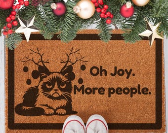 Puede incluir: Una alfombra de coco marrón con un borde negro y un diseño humorístico de temática navideña. Un gato gruñón con cuernos de reno está representado con el texto "Oh Joy. More people."