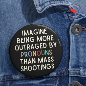 May include: A black round button with the text "IMAGINE BEING MORE OUTRAGED BY PRONOUNS THAN MASS SHOOTINGS" in white and rainbow colors. The button is pinned to a blue denim jacket.