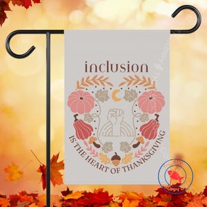 May include: A garden flag with the text "inclusion" and "is the heart of thanksgiving". The flag features a design with pumpkins, leaves, a crescent moon, a fist, and an acorn in shades of pink, orange, and brown.