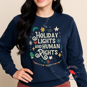 May include: Navy blue sweatshirt with the text "HOLIDAY LIGHTS AND HUMAN RIGHTS" in white and yellow. The design includes festive illustrations of stars, a crescent moon, and a cactus.