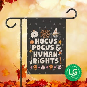 May include: A black rectangular garden flag featuring the white text HOCUS POCUS & HUMAN RIGHTS. The design includes orange and white illustrations of a pumpkin, spider web, spider in a witch hat, stars, candy, a broom, and four small skulls.