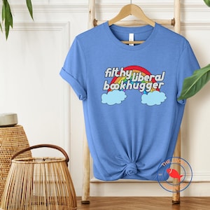 May include: A light blue t-shirt with the text "filthy liberal bookhugger" above a rainbow and clouds design. The shirt is knotted at the waist and hangs on a wooden rack. Wicker baskets are in the foreground.