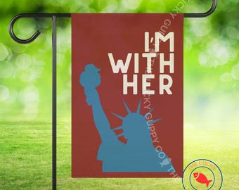 Könnte beinhalten: Eine Gartenflagge mit bordeauxrotem Hintergrund zeigt den Text "I'M WITH HER" in Weiß, mit einer blauen Silhouette der Freiheitsstatue. Die Flagge wird auf einem schwarzen Metallständer vor einem verschwommenen grünen Hintergrund präsentiert.