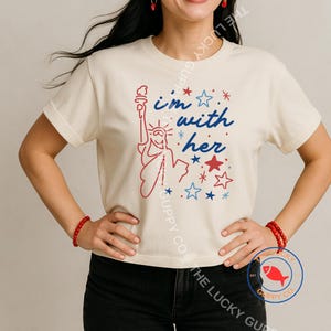 Op de afbeelding: Crèmekleurig T-shirt met een rood en blauw ontwerp van het Vrijheidsbeeld en de tekst "I'm with her", plus sterren. Het shirt heeft korte mouwen en een ronde hals.