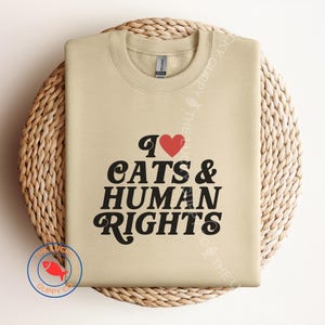 May include: A beige sweatshirt featuring the text "I ❤️ CATS & HUMAN RIGHTS" in black script and a red heart. The garment is presented on a woven, circular placemat. A logo with a red fish is in the lower left corner.