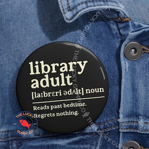May include: A black round button with the text "library adult" in white. The button includes the phonetic pronunciation, definition, and the phrases "Reads past bedtime. Regrets nothing." The button has a red fish logo and the text "The Lucky Guppy Co."