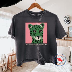 Puede incluir: Una camiseta de manga corta gris carbón con un estampado gráfico de un leopardo verde con manchas negras sobre un fondo cuadrado rosa. Debajo del leopardo, el texto dice "MAY OUR DAUGHTERS BE UNGOVERNABLE" en una fuente sencilla y clara.