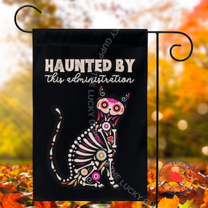 Peut inclure: Un drapeau de jardin noir portant le texte "HAUNTED BY this administration" en blanc et rose clair. Sous le texte se trouve une illustration de squelette de chat stylisé décorée de motifs floraux colorés en rose, rouge, bleu et blanc.