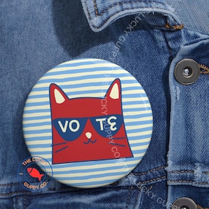 May include: A round button with a red cat wearing sunglasses, with the word "VOTE" in white. The button has a blue and white striped background and the text "LUCKY GUPPY CO" around the edge. The button is on a denim jacket.