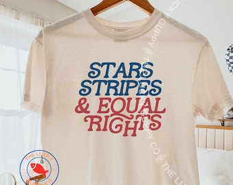 May include: A cream-colored short-sleeve t-shirt featuring the blue text STARS STRIPES and red text & EQUAL RIGHTS in a retro-style font. The shirt is displayed on a wooden hanger against a light-colored background with a circular logo in the corner.