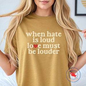 May include: Mustard-colored t-shirt with the text "When hate is loud love must be louder" in a white font. A red heart replaces the word "love". The Lucky Cuppy Co. logo is in the bottom right corner.