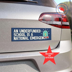 May include: Blue and white bumper sticker with the text "An underfunded school is a national emergency". The sticker has a small graphic of a light blue alarm bell with a yellow outline. The sticker is on the back of a silver car. A red star with the text "Magnet or Sticker" is below the bumper sticker.