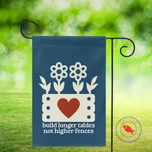 May include: A navy blue garden flag featuring a white floral design and a red heart. The flag displays the text "build longer tables not higher fences" in white. It hangs on a black metal stand.