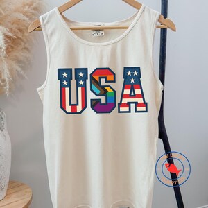 May include: A cream-colored sleeveless tank top featuring the text USA in large block letters. The U and A are decorated with red and white stripes and white stars on a blue background, while the S features a vertical rainbow gradient pattern.