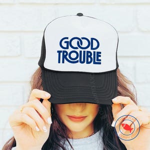 May include: Black and white trucker hat with the words "GOOD TROUBLE" in blue. The hat has a white front panel and a black mesh back. The text is in a bold, sans-serif font.