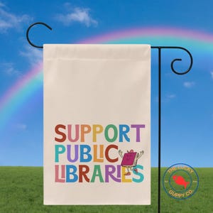 May include: A cream-colored garden flag with the words "SUPPORT PUBLIC LIBRARIES" in colorful letters. A cartoon book character is featured on the right. The flag is hanging on a black metal stand against a blue sky with a rainbow.