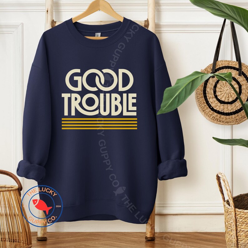 May include: Navy blue sweatshirt with the words "GOOD TROUBLE" in a cream-colored font, with a yellow striped design underneath. The sweatshirt is hanging on a wooden rack.