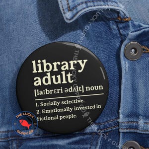 May include: A black round button with the words "library adult" in white, along with a definition. The button also features the text "The Lucky Guppy Co." and a red fish logo. The button is set against a denim jacket.