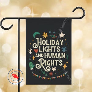 May include: A black rectangular garden flag featuring the text "HOLIDAY LIGHTS AND HUMAN RIGHTS" in cream-colored lettering. The design includes colorful illustrations of stars, a crescent moon, a heart, a mitten, and festive bunting.