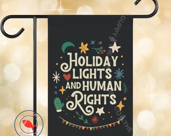 Può includere: Una bandiera da giardino nera con la scritta bianca "HOLIDAY LIGHTS AND HUMAN RIGHTS", circondata da illustrazioni colorate di stelle, una luna crescente e un cactus. La bandiera è esposta su un supporto in metallo nero.
