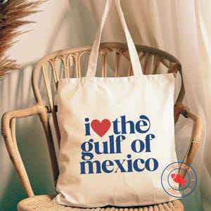 May include: A white canvas tote bag with blue text that reads "i ♥ the gulf of mexico" and a red heart. The bag has a small logo in the bottom right corner that reads "The Lucky Goppy Co."