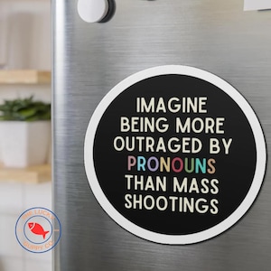 May include: A round black and white magnet with the text "Imagine being more outraged by pronouns than mass shootings" in colorful letters.