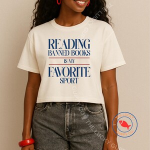 May include: Cream-colored crop top with the text "READING BANNED BOOKS IS MY FAVORITE SPORT" in navy blue. The text is separated by red lines. The shirt is paired with denim jeans.
