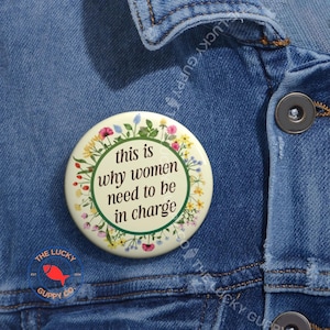 May include: A round, cream-colored button with the text "this is why women need to be in charge" in a green circle, surrounded by a floral wreath. The button is pinned to a blue denim jacket.