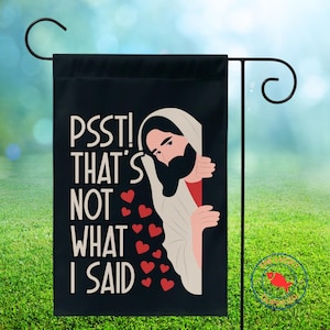 May include: Black garden flag with the text "PSST! THAT'S NOT WHAT I SAID" and an illustration of Jesus peeking out. Red hearts are scattered around the illustration. The flag is displayed on a black metal garden stake.