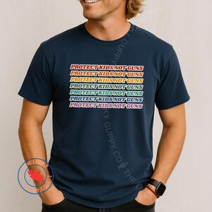 May include: Navy blue t-shirt with the text "PROTECT KIDS NOT GUNS" in a rainbow gradient design. The text is repeated multiple times across the front of the shirt. The shirt is a short-sleeved crew neck.