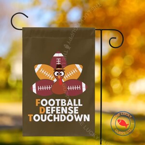 Könnte beinhalten: Eine braune Gartenflagge mit einem Truthahn-Design aus American-Football-Bällen. Die Flagge trägt die weißen Worte "FOOTBALL DEFENSE TOUCHDOWN". Der Truthahn trägt einen Zylinder. Der Hintergrund ist verschwommen.