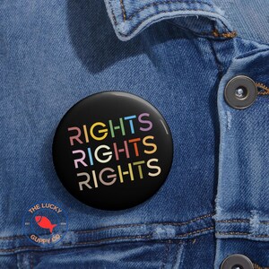May include: A black badge with the word "RIGHTS" in rainbow colours repeated three times. The badge is pinned to a blue denim jacket. The Lucky Guppy Co. logo is visible.