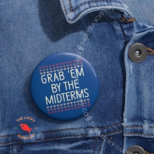 May include: A round, blue button with the text "GRAB 'EM BY THE MIDTERMS" in white, surrounded by red and white stars. The button is on a denim jacket. The Lucky Guppy Co. logo is visible.